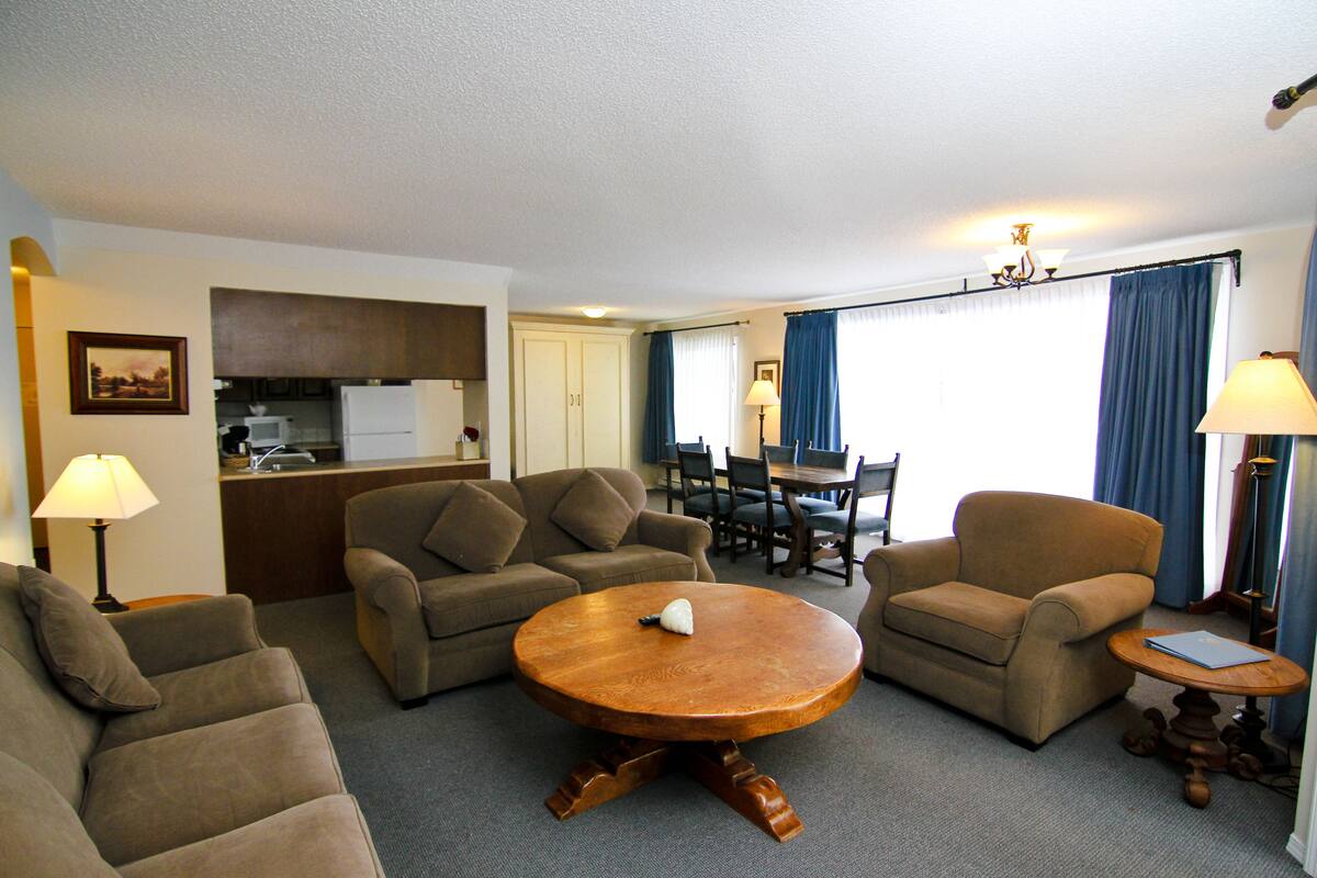 Habitación de hotel con sala de estar que incluye un conjunto de sofás, una mesa de centro de madera y una mesa de comedor con sillas.