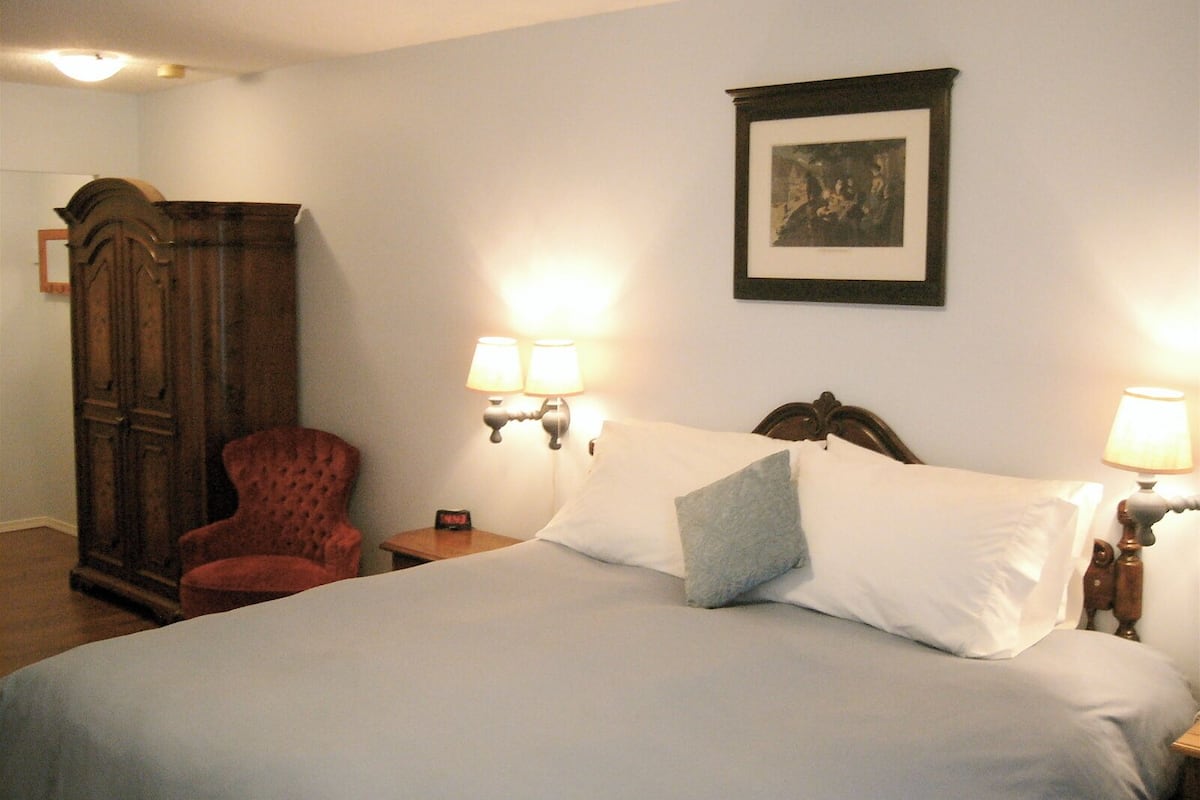 Habitación de hotel con una cama grande, un armario de madera, un sillón rojo, dos lámparas de pared y un cuadro en la pared.