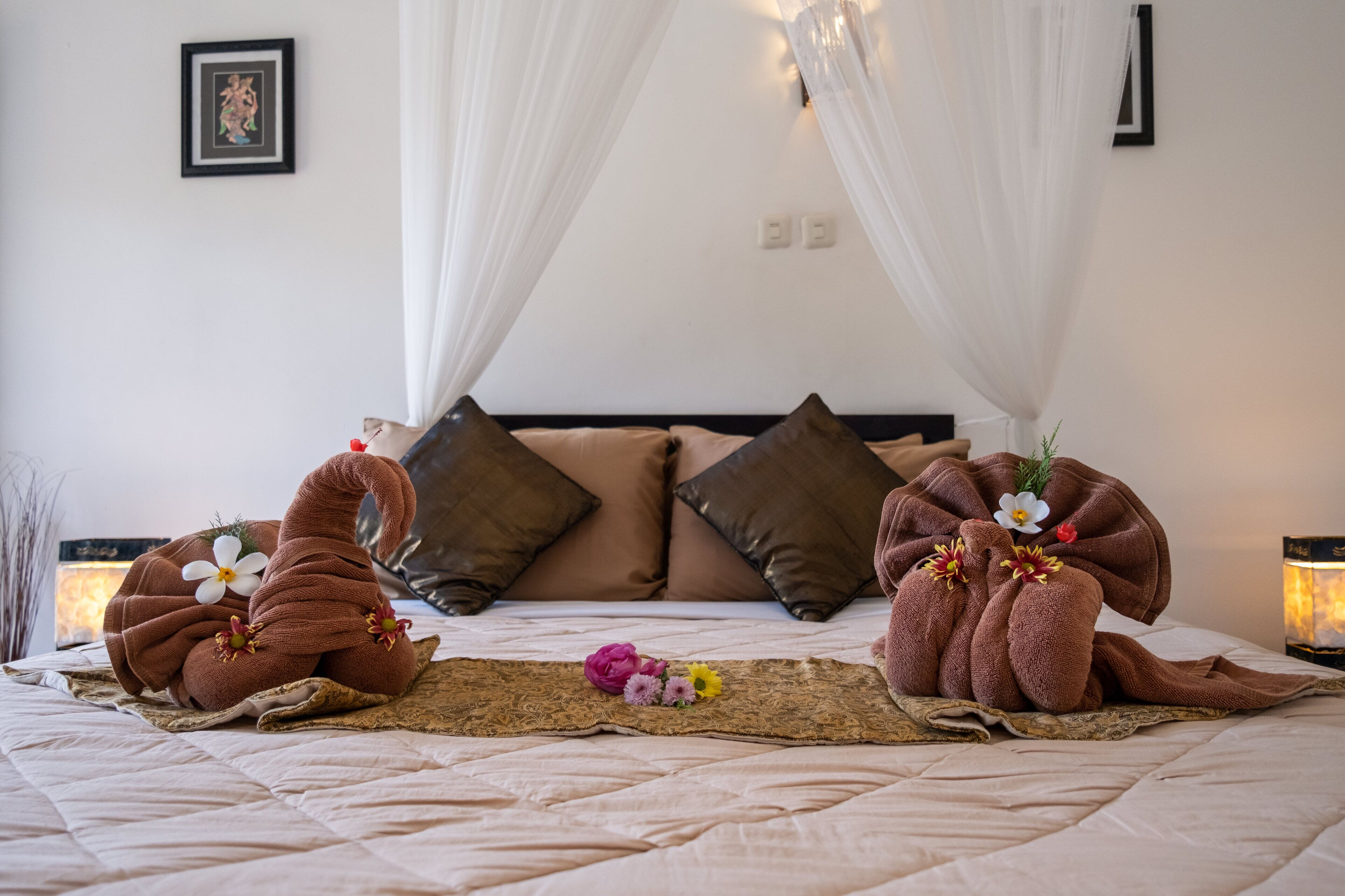 A neatly made bed with towels arranged in swan shapes, a decorative pillow, and a small floral arrangement on a woven mat.