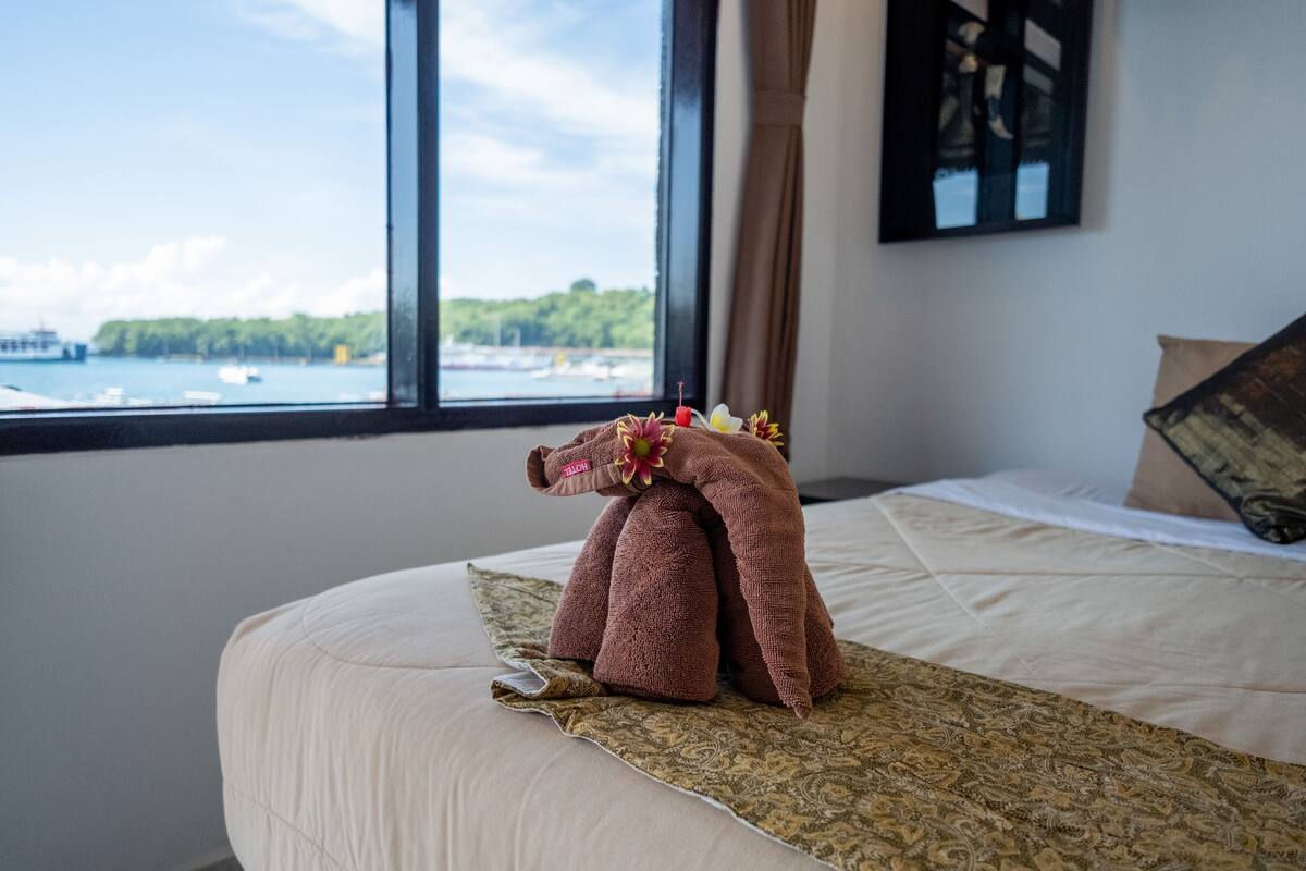 A bed with a patterned bedspread, a brown towel shaped like an elephant, and a window with a view of boats and greenery.