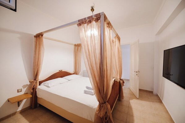 A four-poster bed with sheer curtains, a wooden bedside table, and a flat-screen TV mounted on the wall.