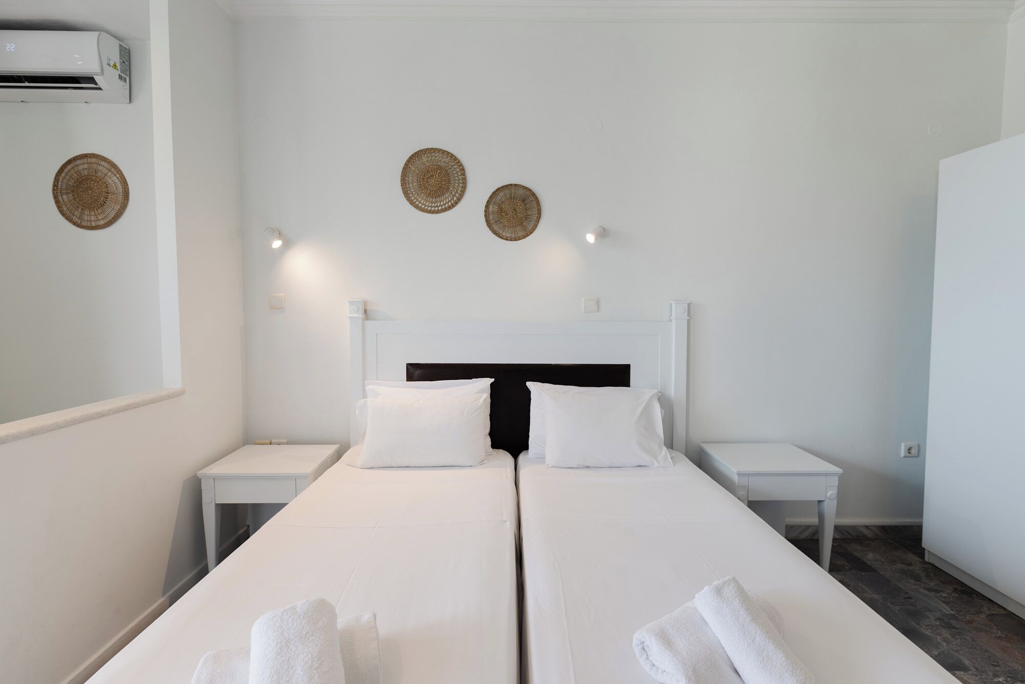 Habitación de hotel con dos camas, ropa de cama blanca, cabecero blanco y aire acondicionado de pared.
