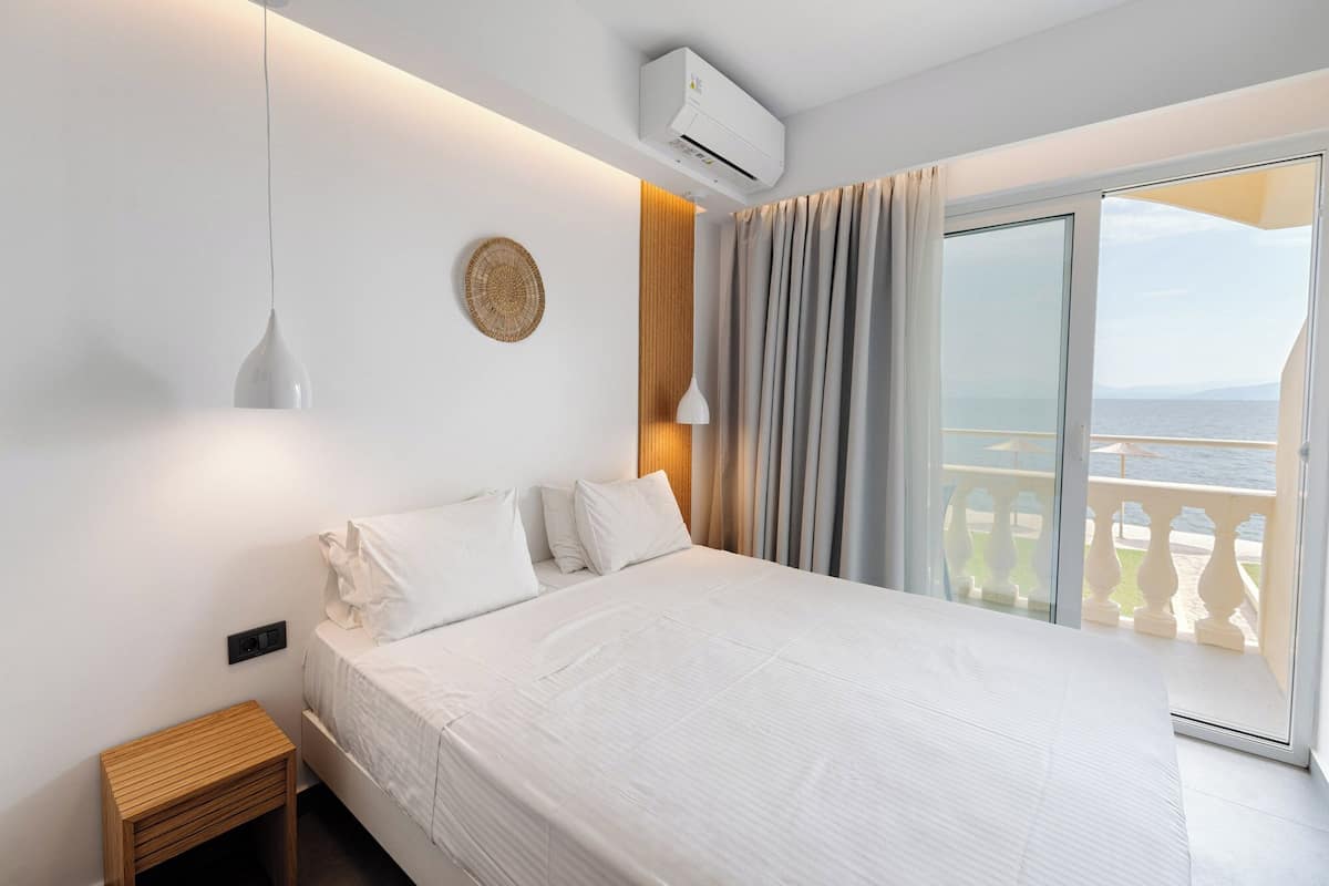 Un dormitorio con cama, mesita de noche, equipo de aire acondicionado, lámpara de pared y vista al mar a través de una puerta corrediza de vidrio.