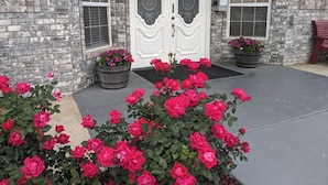 A stone building with a white door marked "3454B" and two potted plants with pink flowers in front.