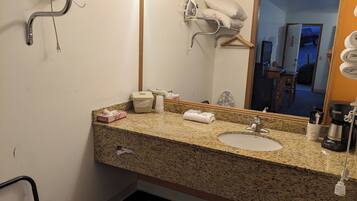 A hotel bathroom with a granite countertop, a sink, a mirror, a towel rack, and a shower area.