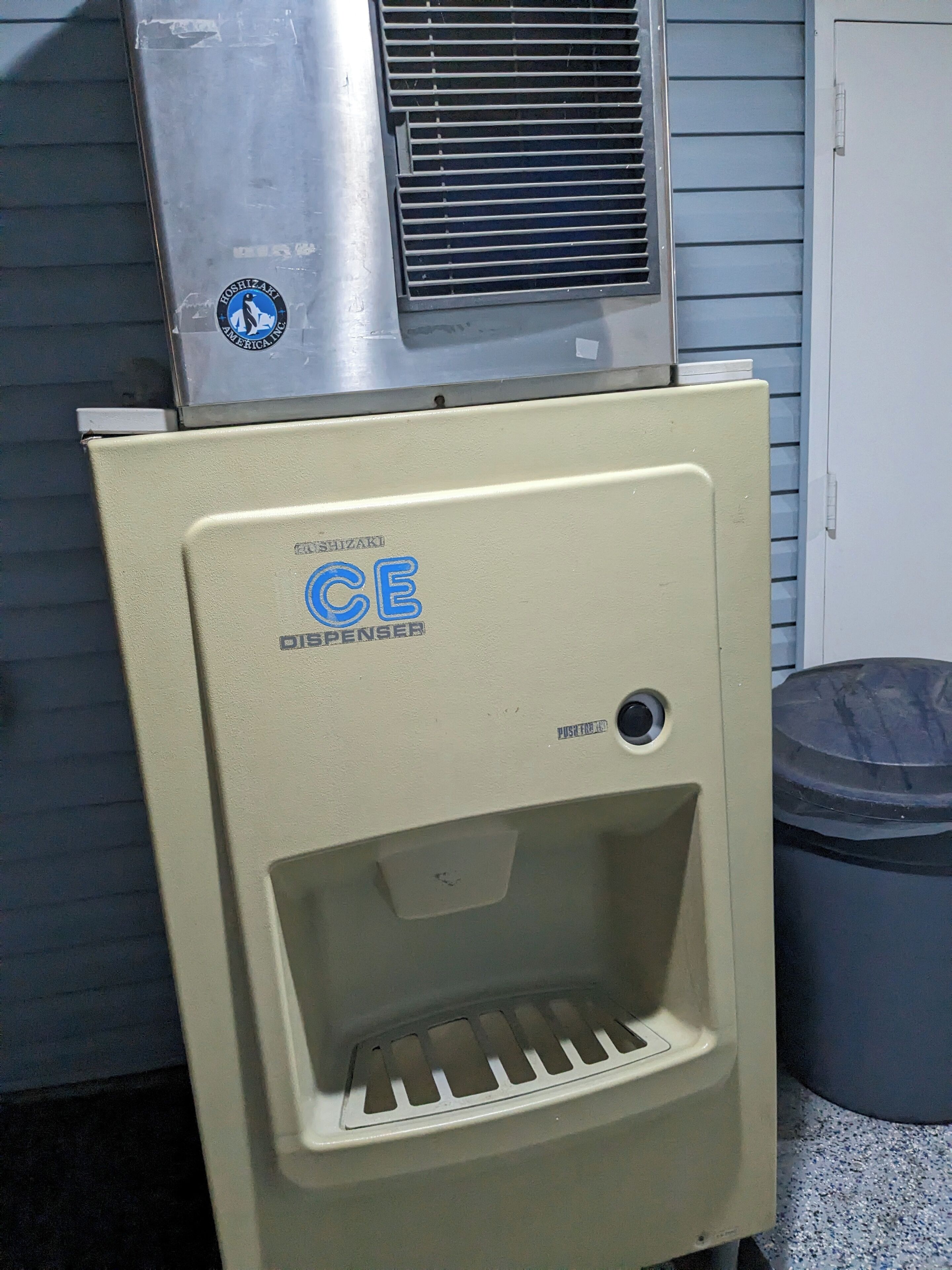 Un distributeur automatique de couleur beige, équipé d’un point d’eau et situé à proximité d’une poubelle.