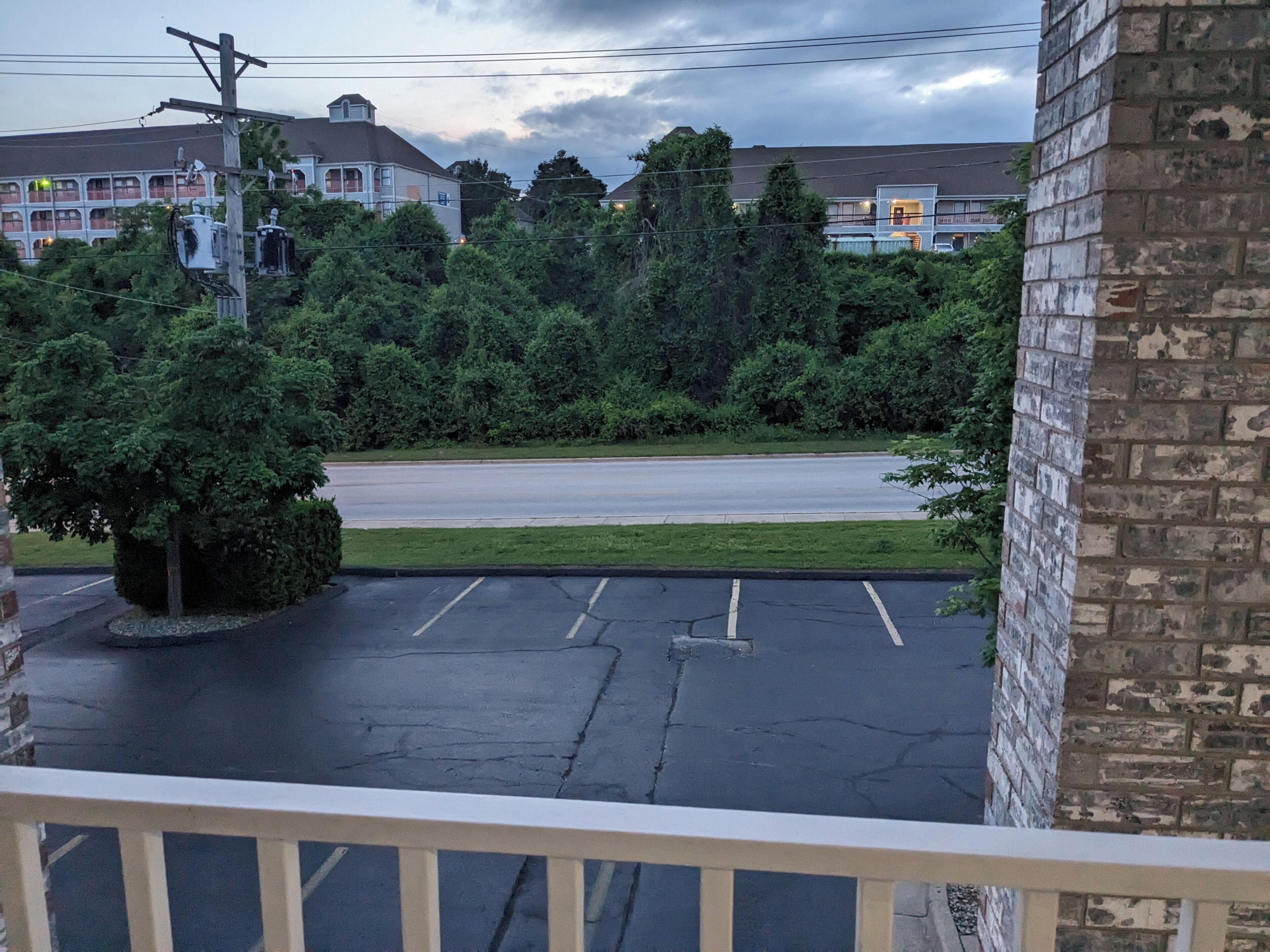 A view from a balcony overlooking a parking lot with a road, trees, and buildings in the background.