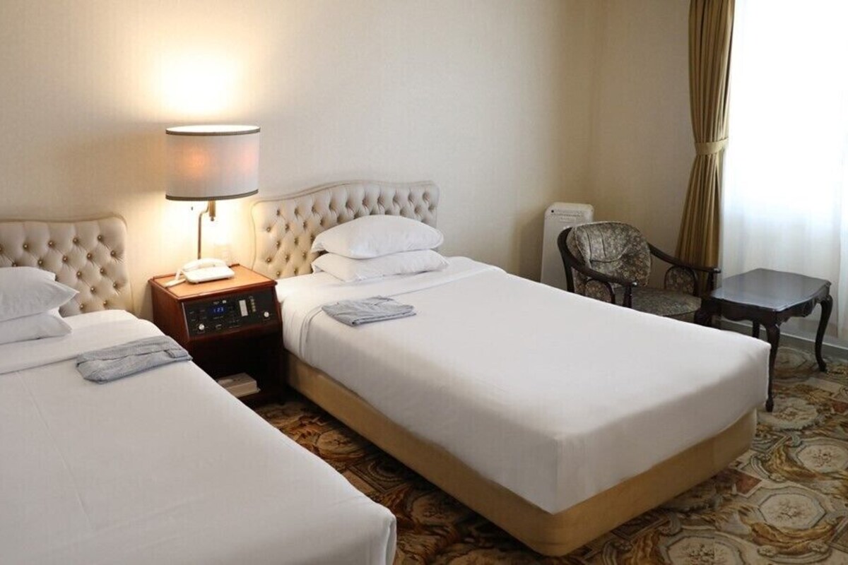 A hotel room with two beds, a bedside table with a lamp, a chair, and a small table.