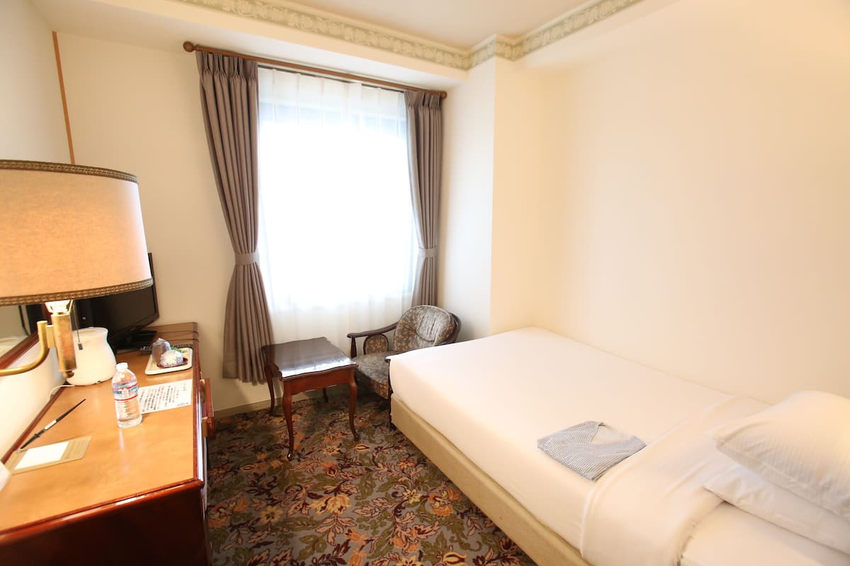 A hotel room with a bed, a desk with a lamp, a chair, a small table, and a window with curtains.