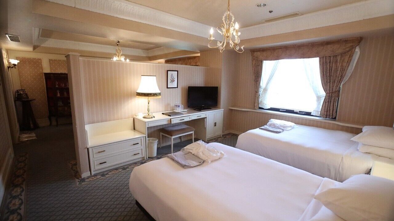 A hotel room with two beds, a desk with a television, a chandelier, and a window with curtains.
