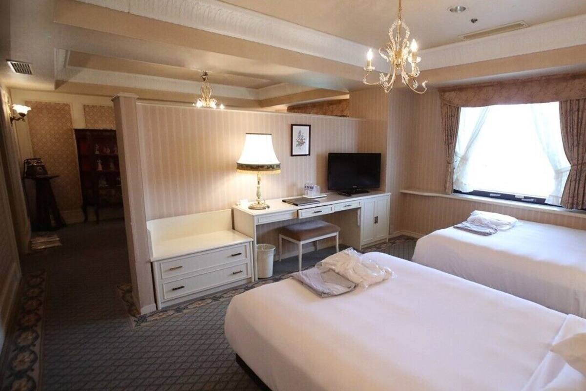 A hotel room with two beds, a desk with a television, a chandelier, and a window with curtains.