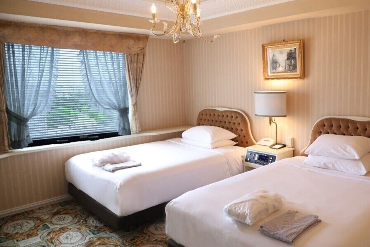 A hotel room with two beds, a chandelier, a bedside table with a lamp, a painting on the wall, and a window with curtains.