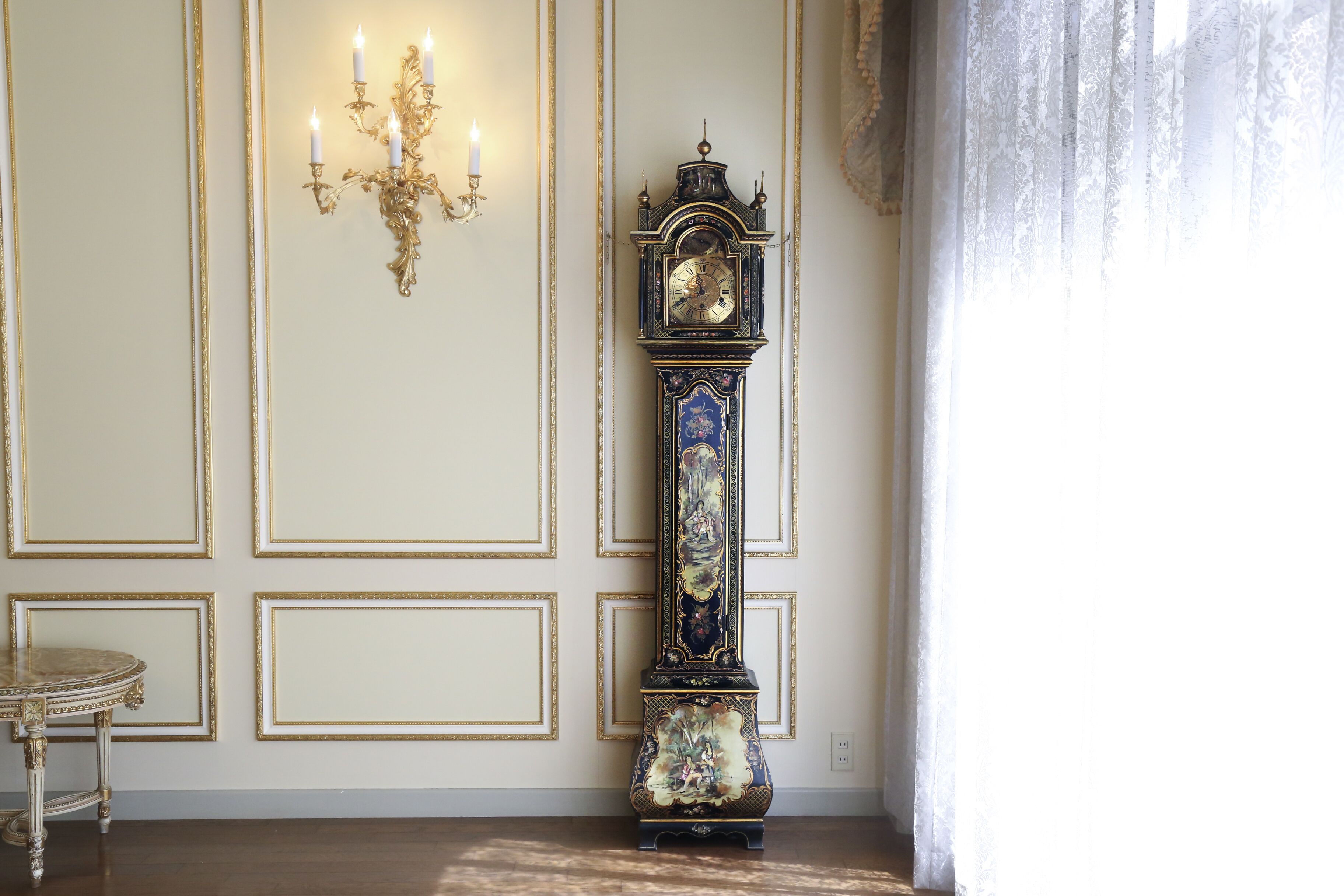 A grand room with a tall, ornate grandfather clock, a marble table, and a wall-mounted candelabra.