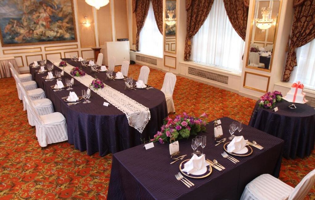 Un salón de banquetes con largas mesas dispuestas para un evento formal, con sillas blancas, manteles oscuros y centros de mesa florales.