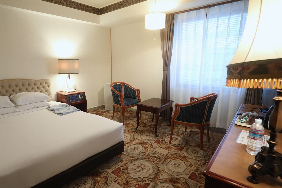 A hotel room with a bed, two chairs, a desk, a lamp, a TV, and a water bottle.