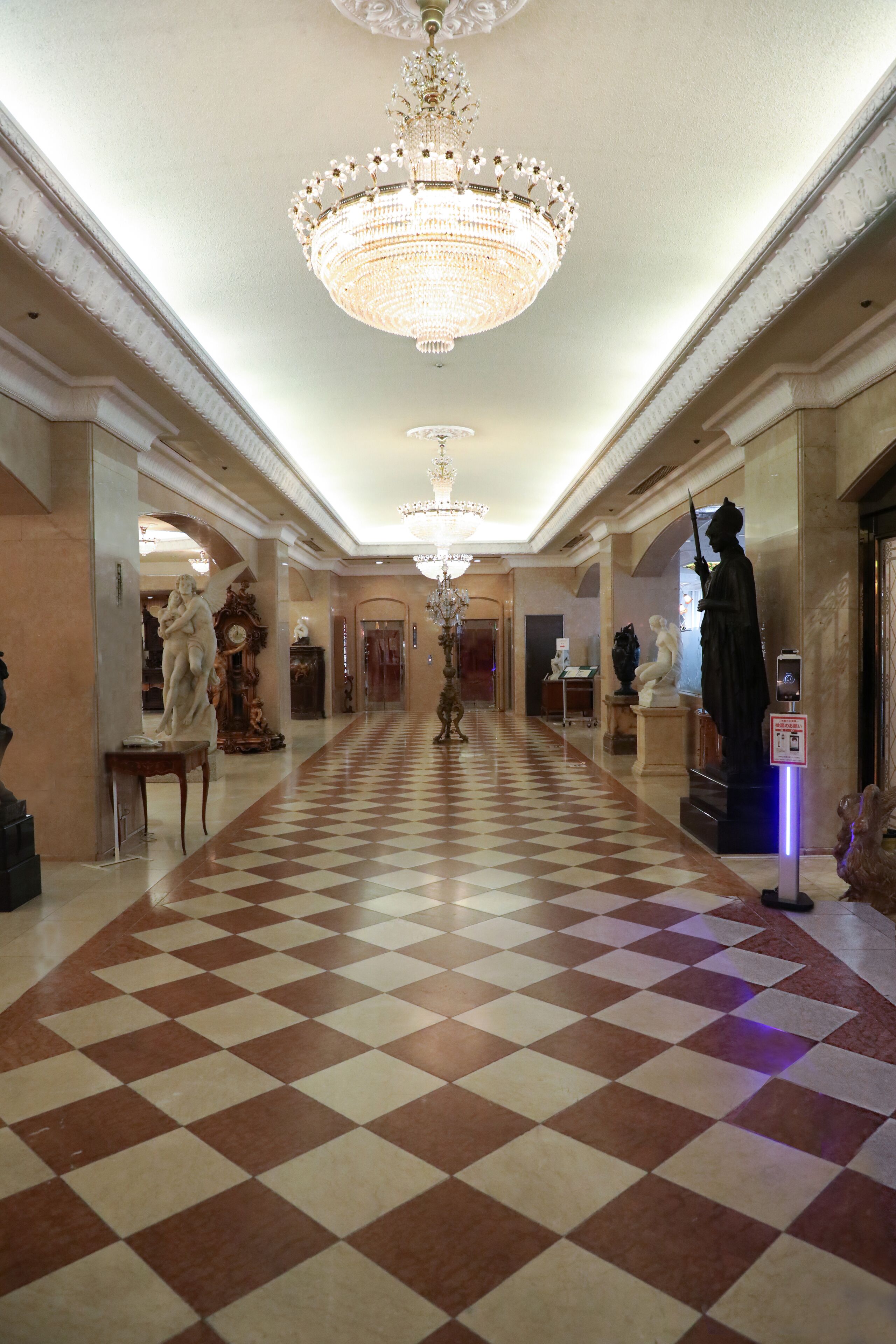 A grand hallway with a checkered floor, ornate chandeliers, and classical statues.