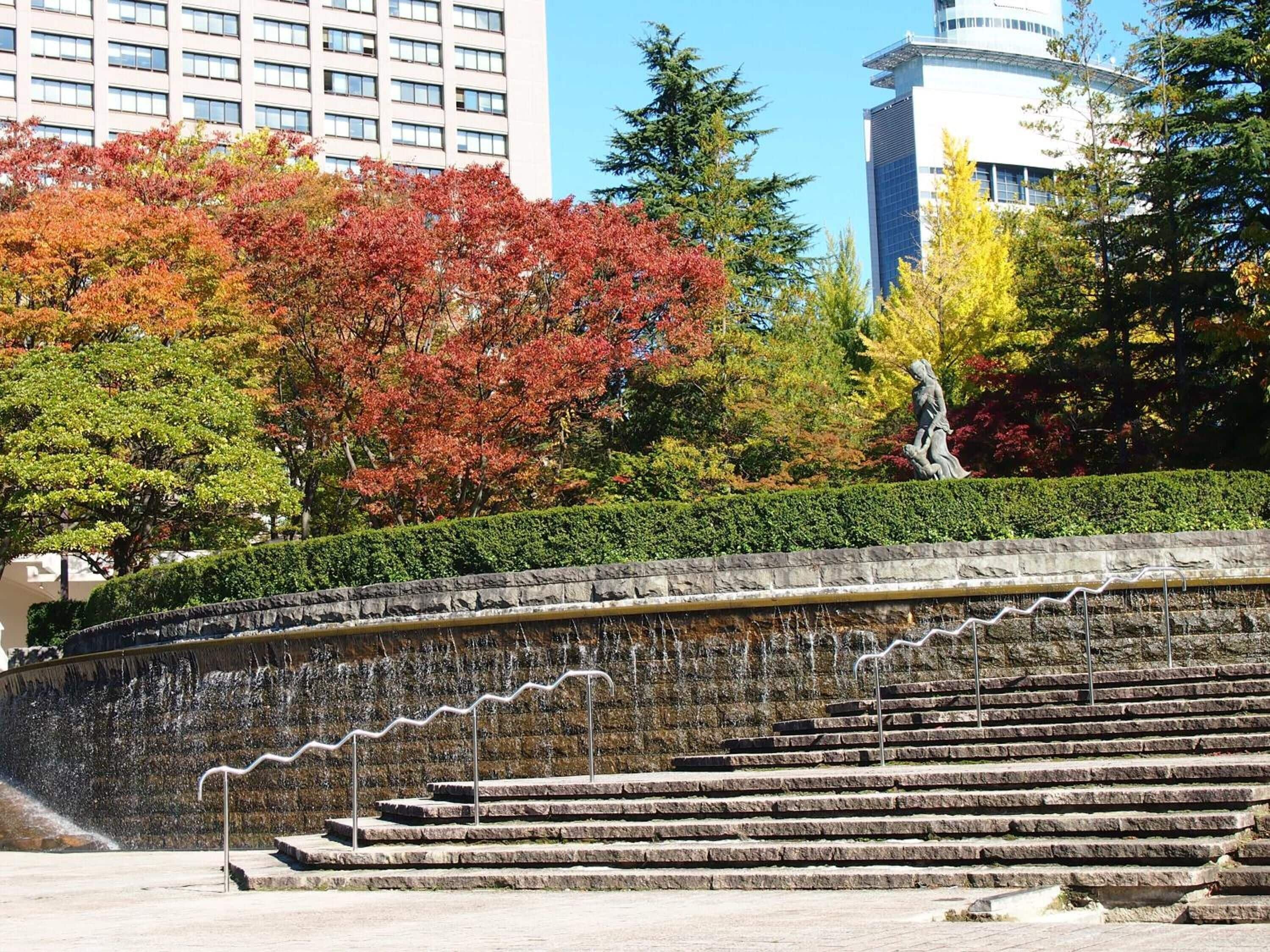 Una escalera de piedra conduce a una plataforma de observación, rodeada de árboles con follaje otoñal y un edificio moderno al fondo.