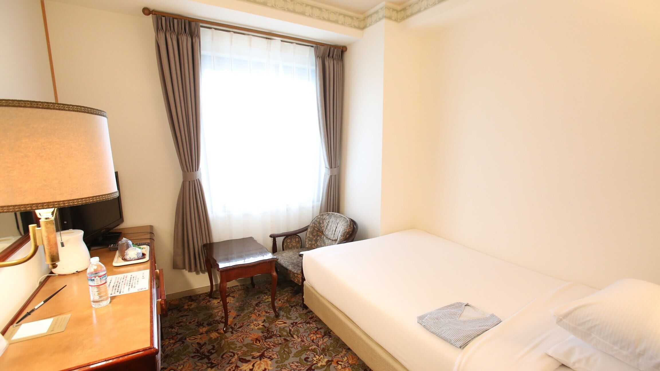A hotel room with a bed, a desk with a lamp, a chair, a window with curtains, and a carpeted floor.