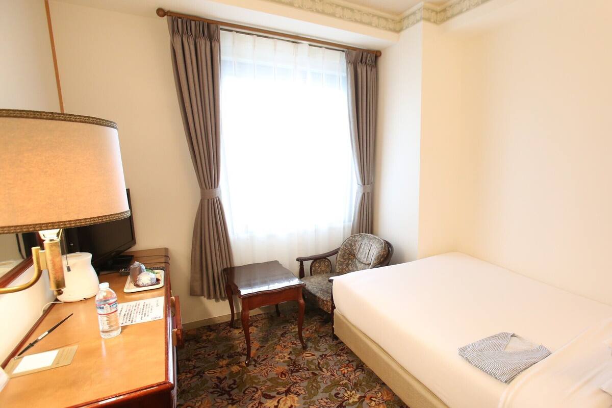A hotel room with a bed, a desk with a lamp, a chair, a window with curtains, and a carpeted floor.