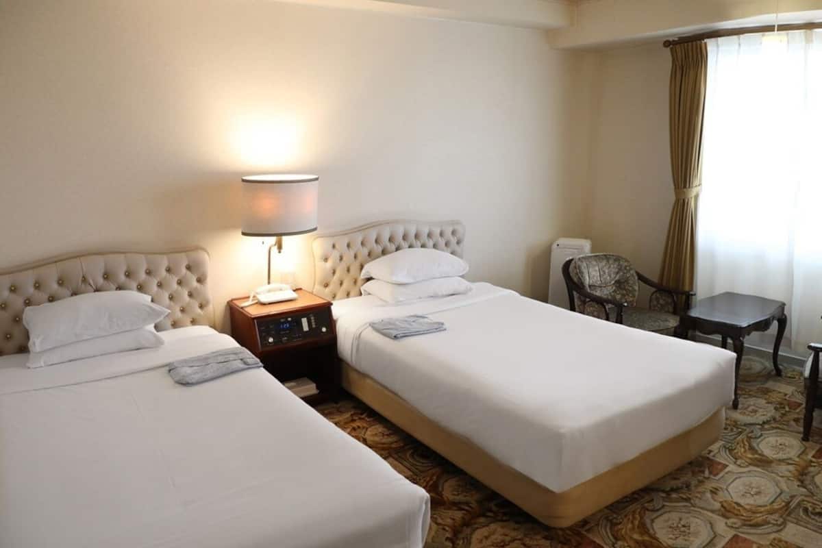 A hotel room with two beds, a bedside table with a lamp, a chair, and a small table.