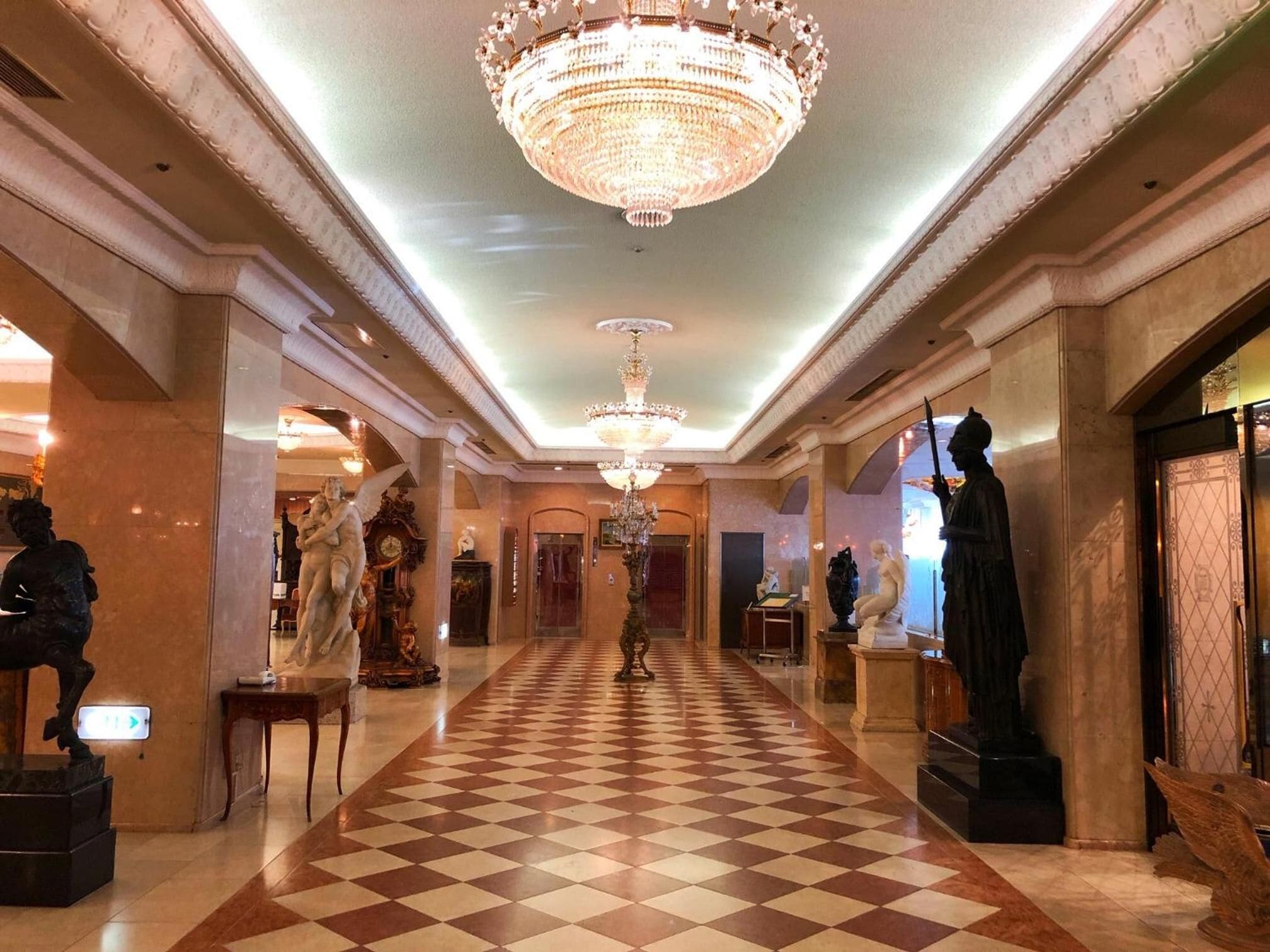 A grand hallway with a checkered floor, a large chandelier, and statues along the walls.