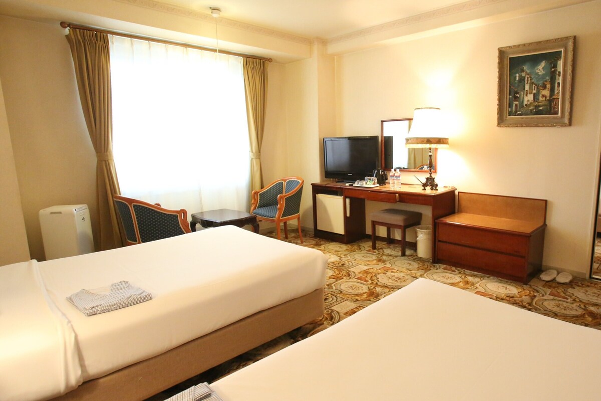 A hotel room with two beds, a desk with a television, a chair, a lamp, and a painting on the wall.