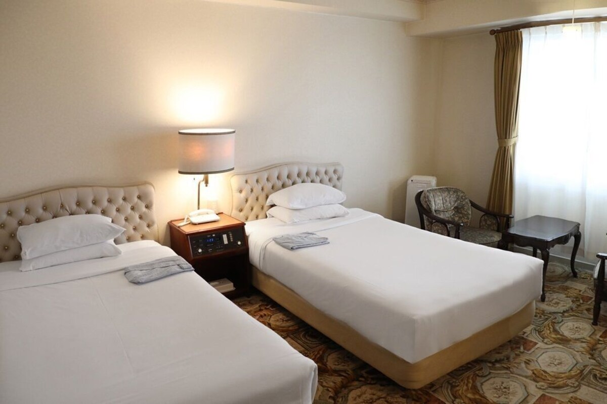 A hotel room with two beds, a bedside table with a lamp, a chair, and a small table.