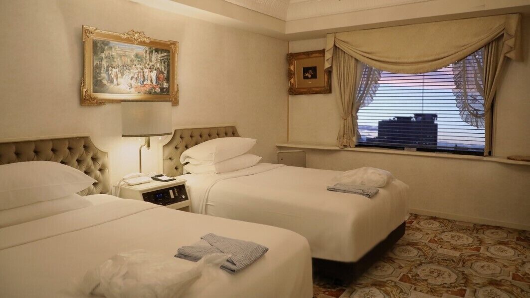 A hotel room with two beds, a bedside table with a lamp, a picture frame, a window with curtains, and a patterned floor.