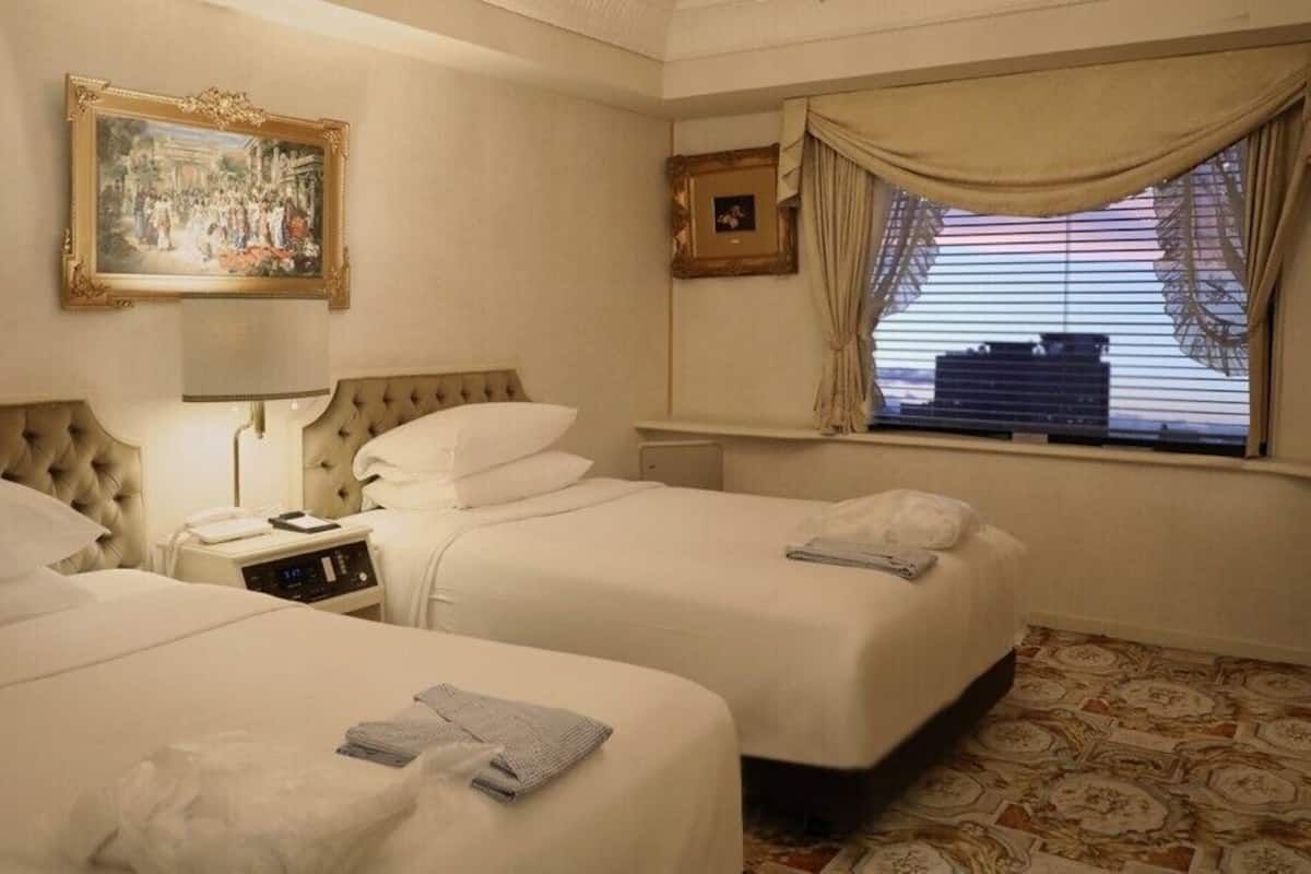 A hotel room with two beds, a bedside table with a lamp, a picture frame, a window with curtains, and a patterned floor.