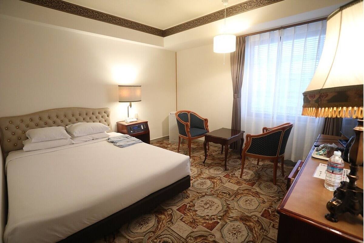A hotel room with a large bed, a desk, two chairs, a lamp, and a window with curtains.