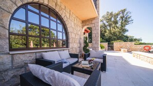 Fachada de piedra con un gran ventanal arqueado y zona de estar exterior con mobiliario de mimbre y cojines.
