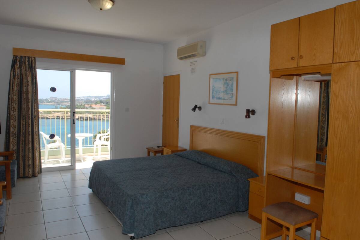 A bedroom with a bed, a wooden wardrobe, a small table, and a chair. There is a sliding glass door leading to a balcony with a view of the sea.