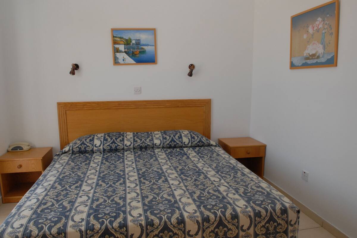 A bedroom with a wooden headboard, a patterned bedspread, two bedside tables, and two wall-mounted paintings.