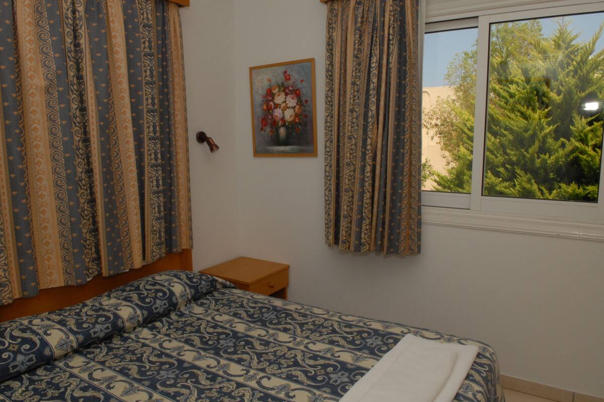 A bedroom with a patterned bedspread, a small wooden nightstand, a framed floral painting on the wall, and a window with a view of trees.