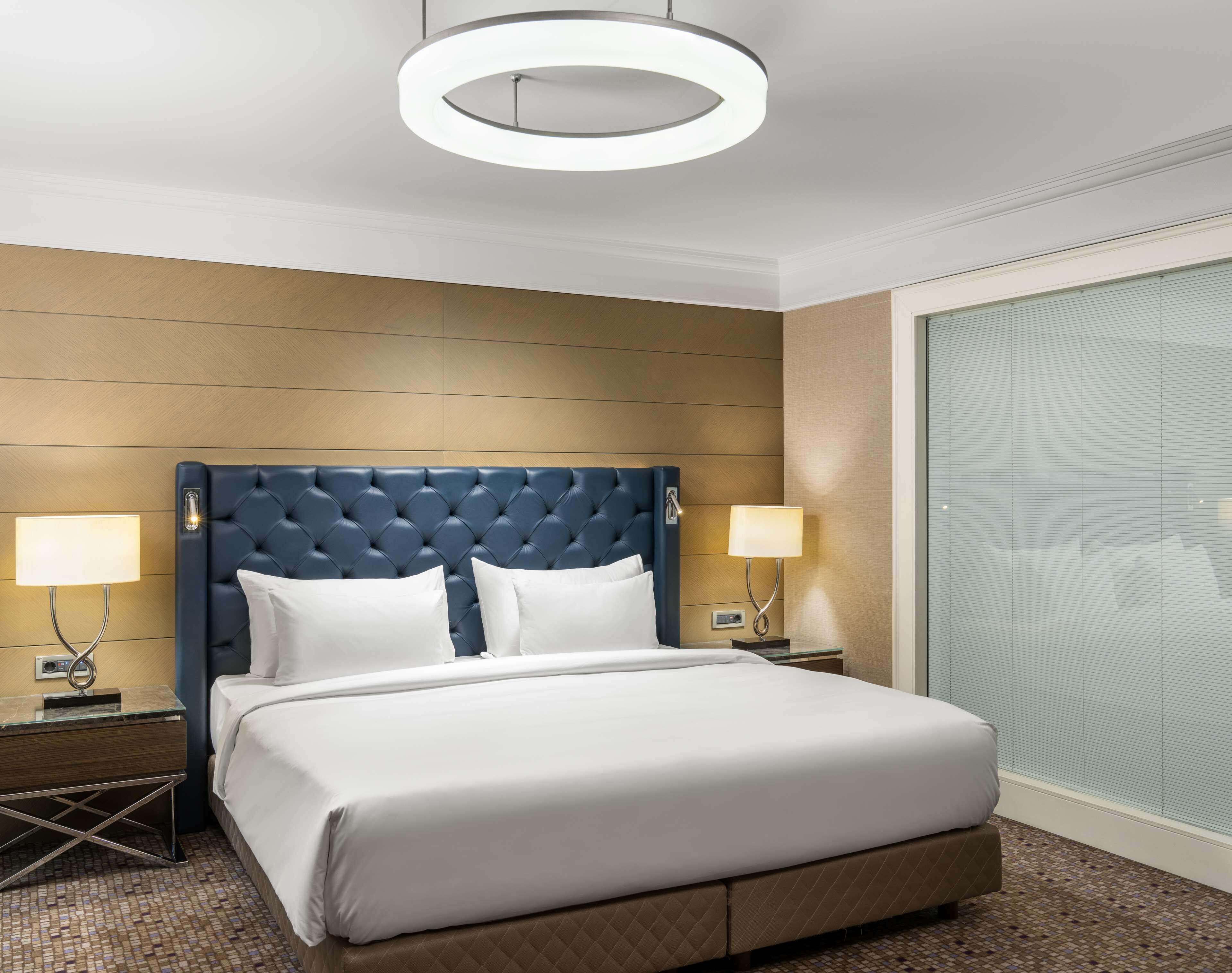 Une chambre d’hôtel avec un grand lit, deux lampes de chevet, un lustre circulaire moderne et une grande fenêtre avec des stores.