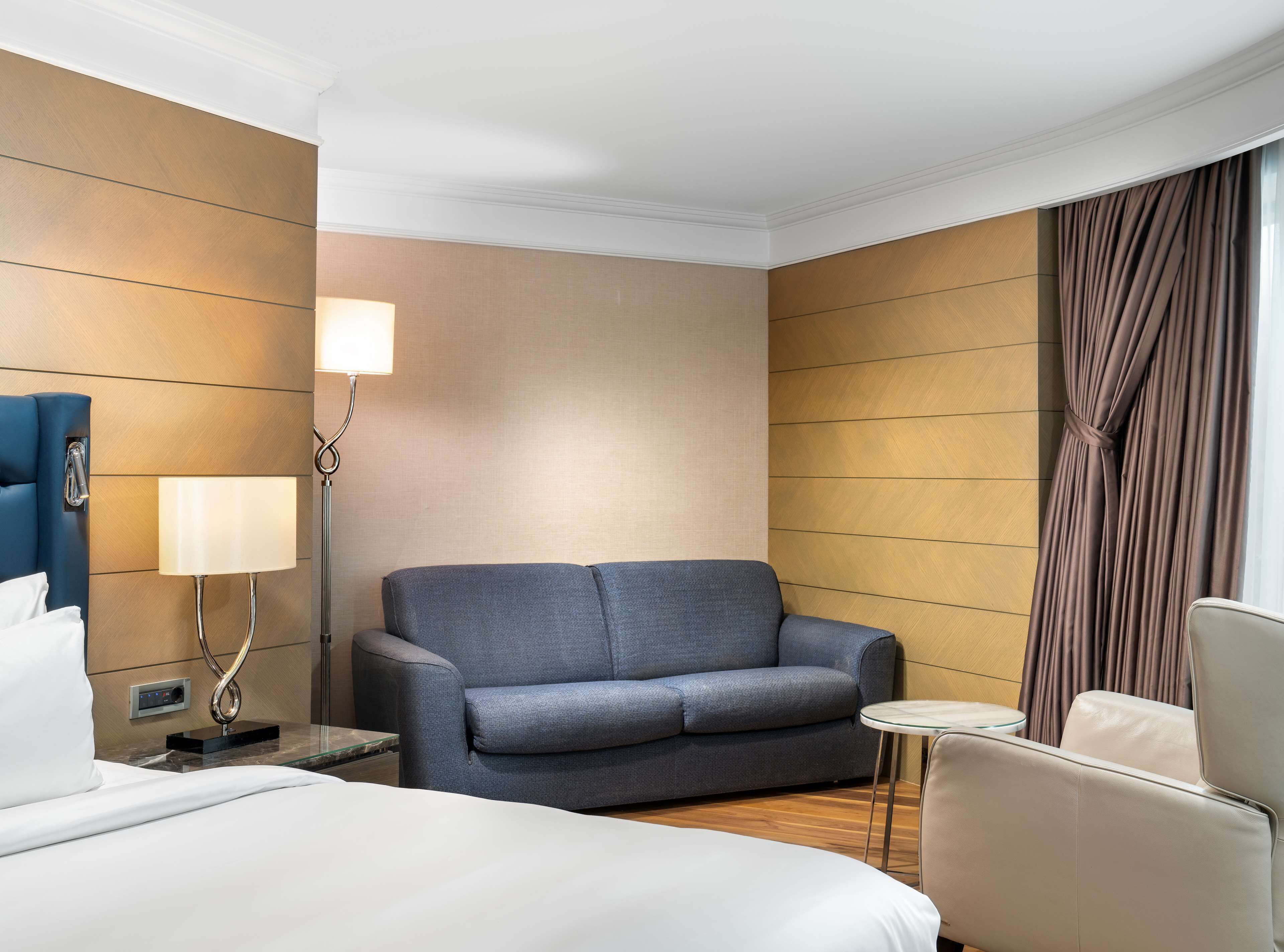 Une chambre d’hôtel avec un lit, un canapé gris, une petite table ronde, une lampe et un rideau.