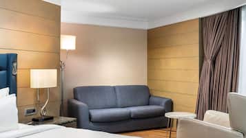 Une chambre d’hôtel avec un lit, un canapé gris, une petite table ronde, une lampe et un rideau.