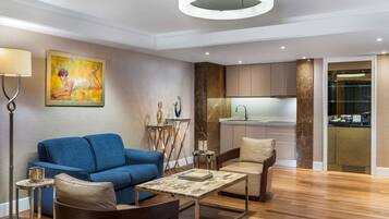 A modern living room with a blue sofa, a coffee table, and a chair. There is a painting on the wall, a floor lamp, and a kitchen area visible through an open door.