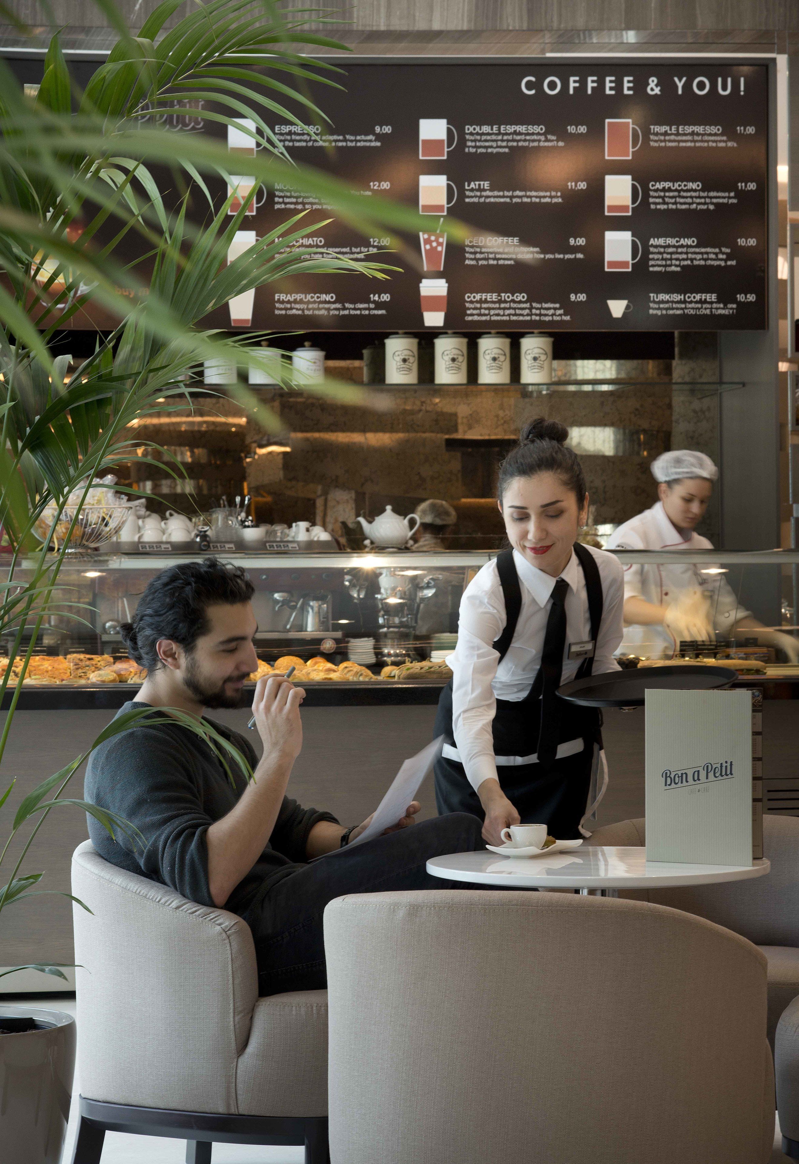 A coffee shop interior with a menu board, a waiter serving a customer, and a display case in the background.