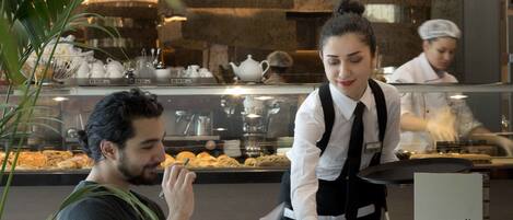 A coffee shop interior with a menu board, a waiter serving a customer, and a display case in the background.