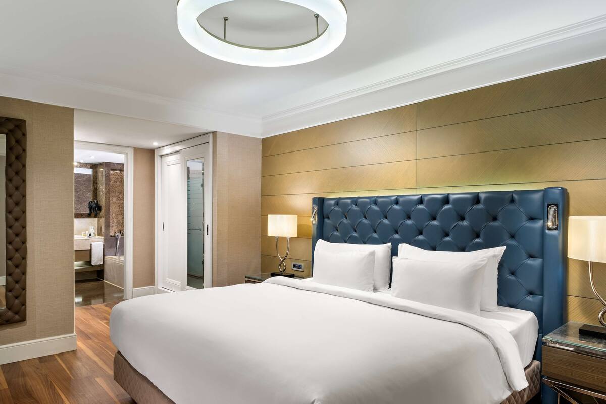 A modern hotel room with a large bed, a tufted headboard, a round ceiling light, and a wooden panel wall.