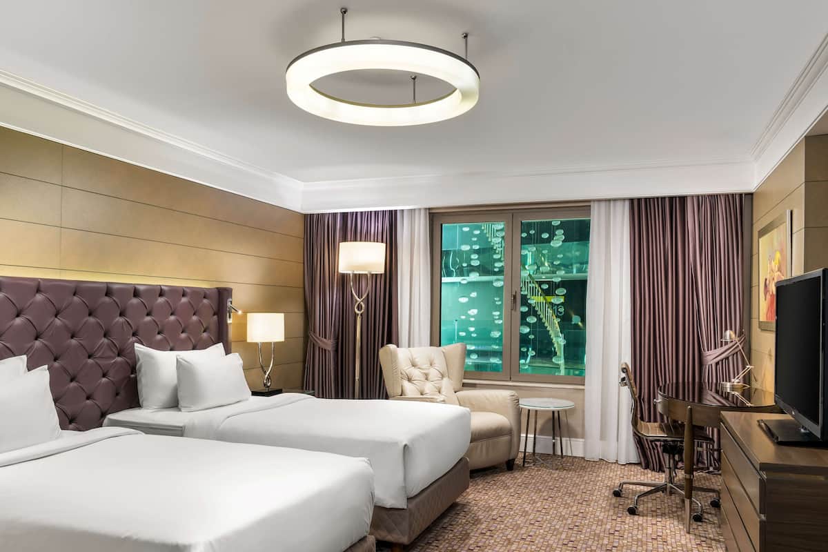 A hotel room with two beds, a desk, a chair, a television, and a large window with curtains.