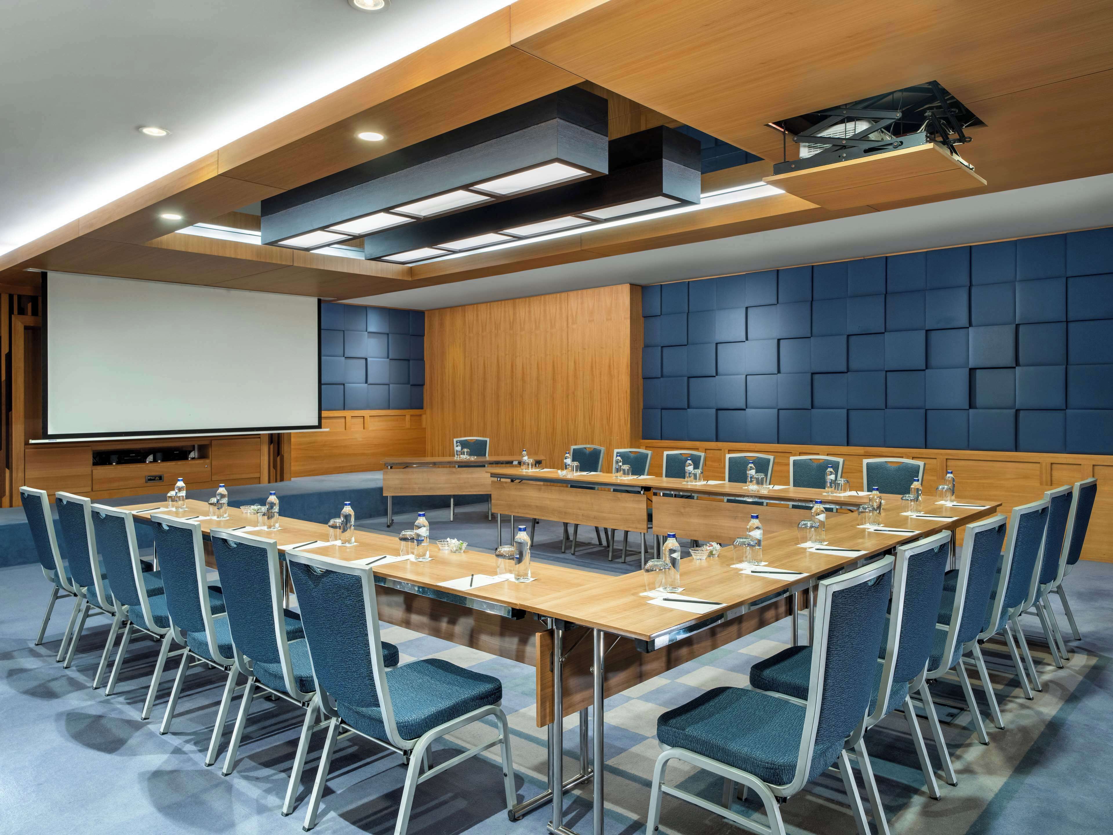 A conference room with a long table, blue chairs, and a projector screen.