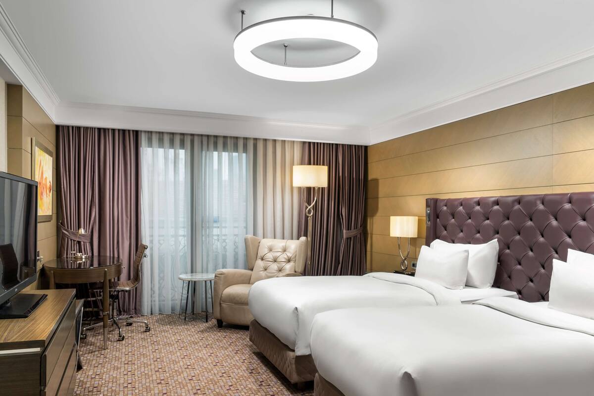 A hotel room with a large bed, a desk with a chair, a television, and a view of the city through the window.