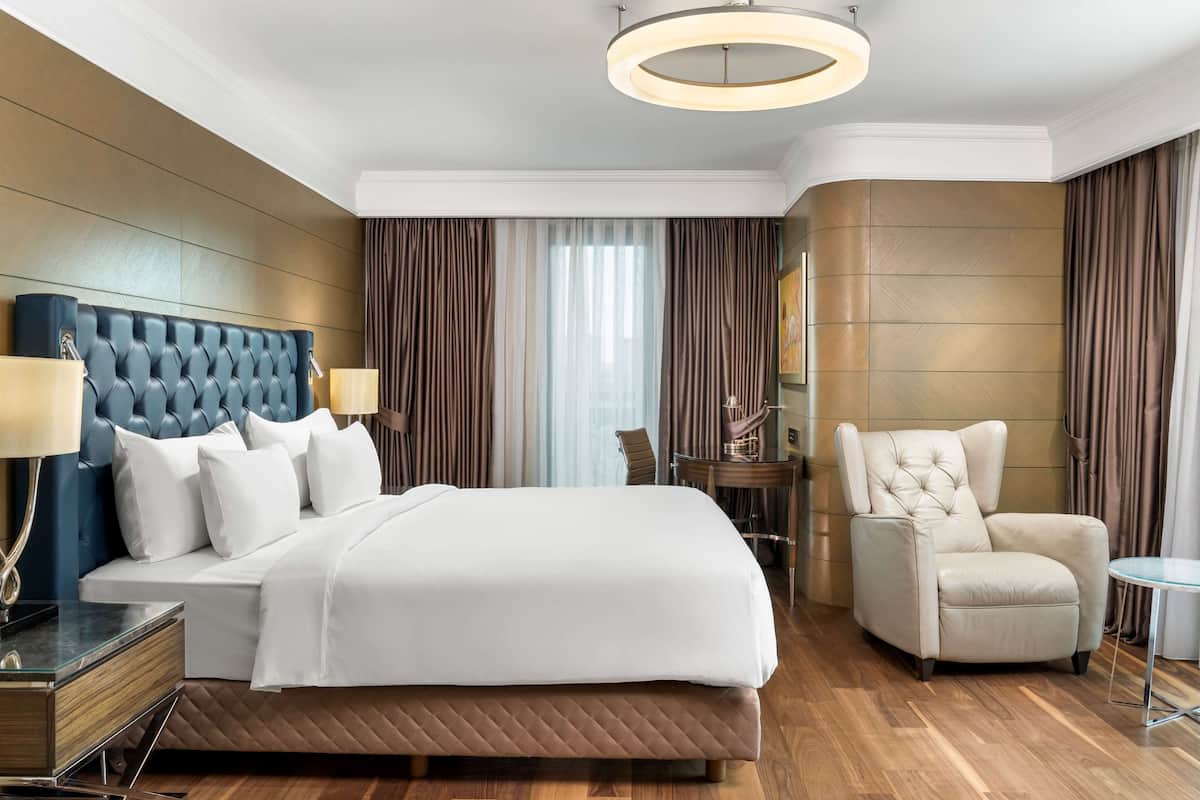 A modern hotel room with a large bed, a sitting area with a chair, a desk, and a round ceiling light.