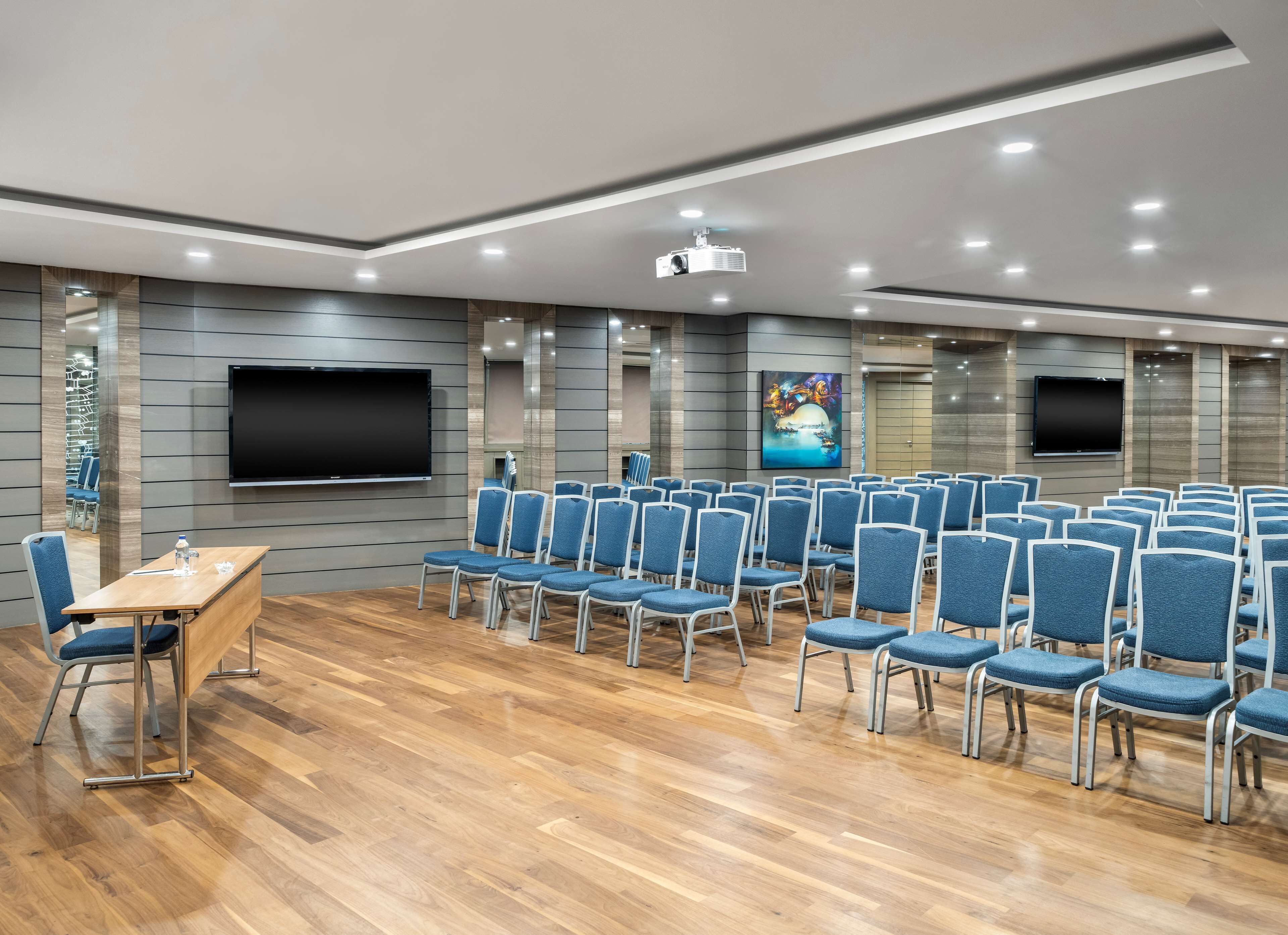 A conference room with rows of blue chairs, a wooden table, and two large flat-screen TVs mounted on the walls.