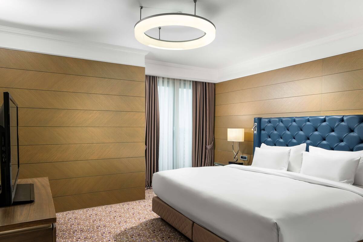 A hotel room with a large bed, a television on a wooden nightstand, a wall-mounted lamp, and a window with curtains.