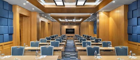A conference room with rows of tables and chairs, a large screen at the front, and wooden paneling on the walls.