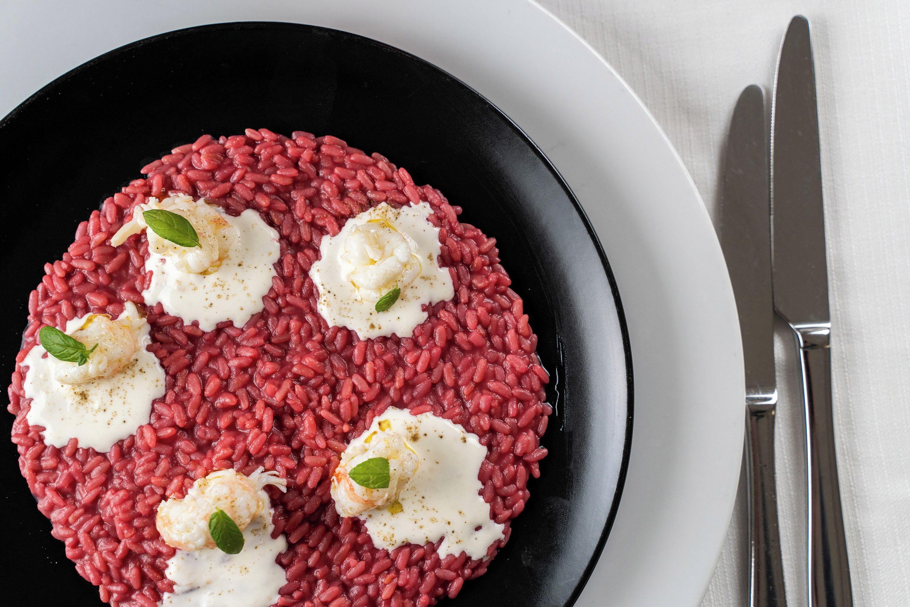 Un plato negro con risotto rojo y cucharadas de crema, adornado con hojas de albahaca.