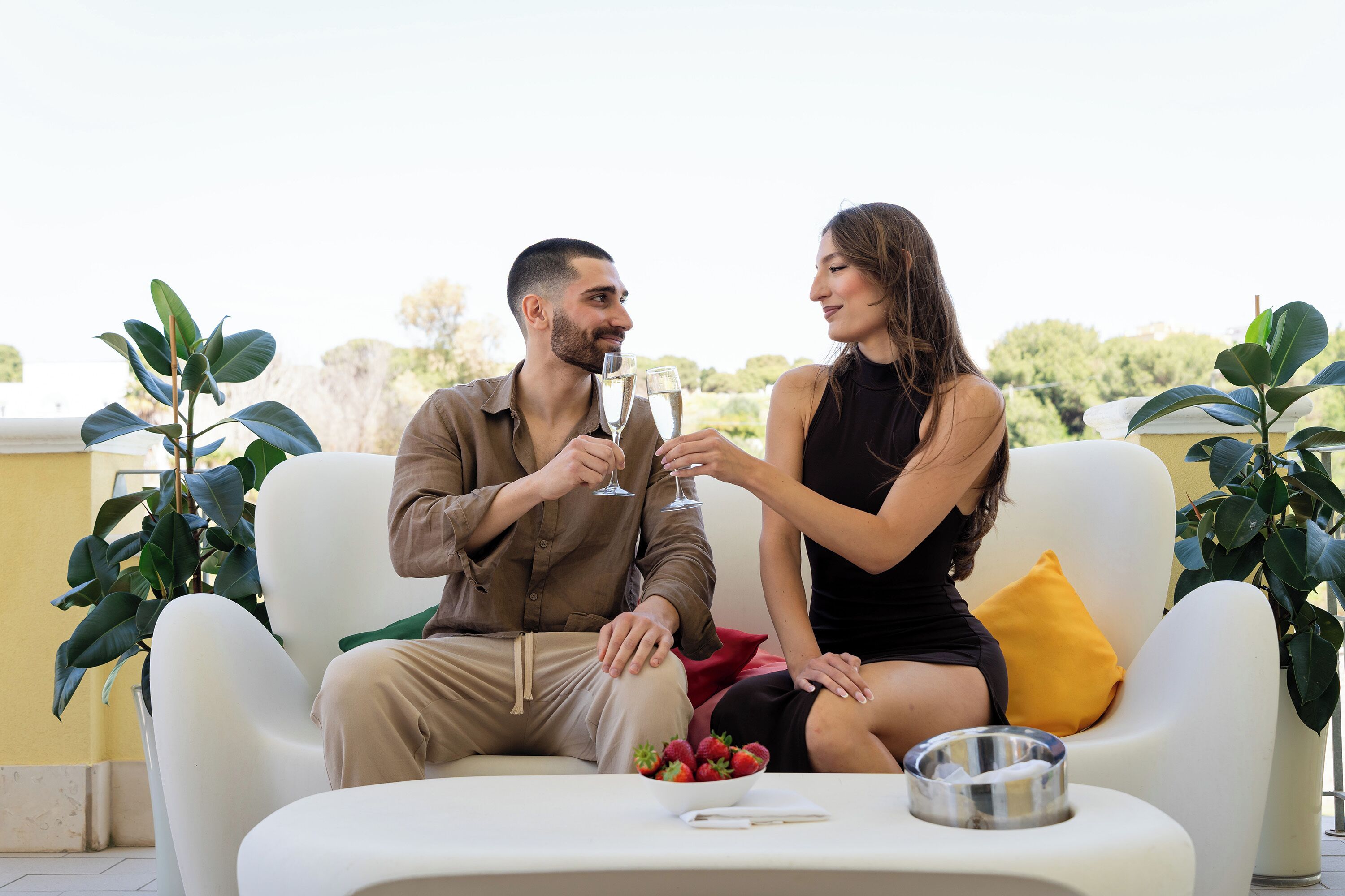 A couple sitting on a white sofa with a bowl of strawberries and a metal ashtray on a table.